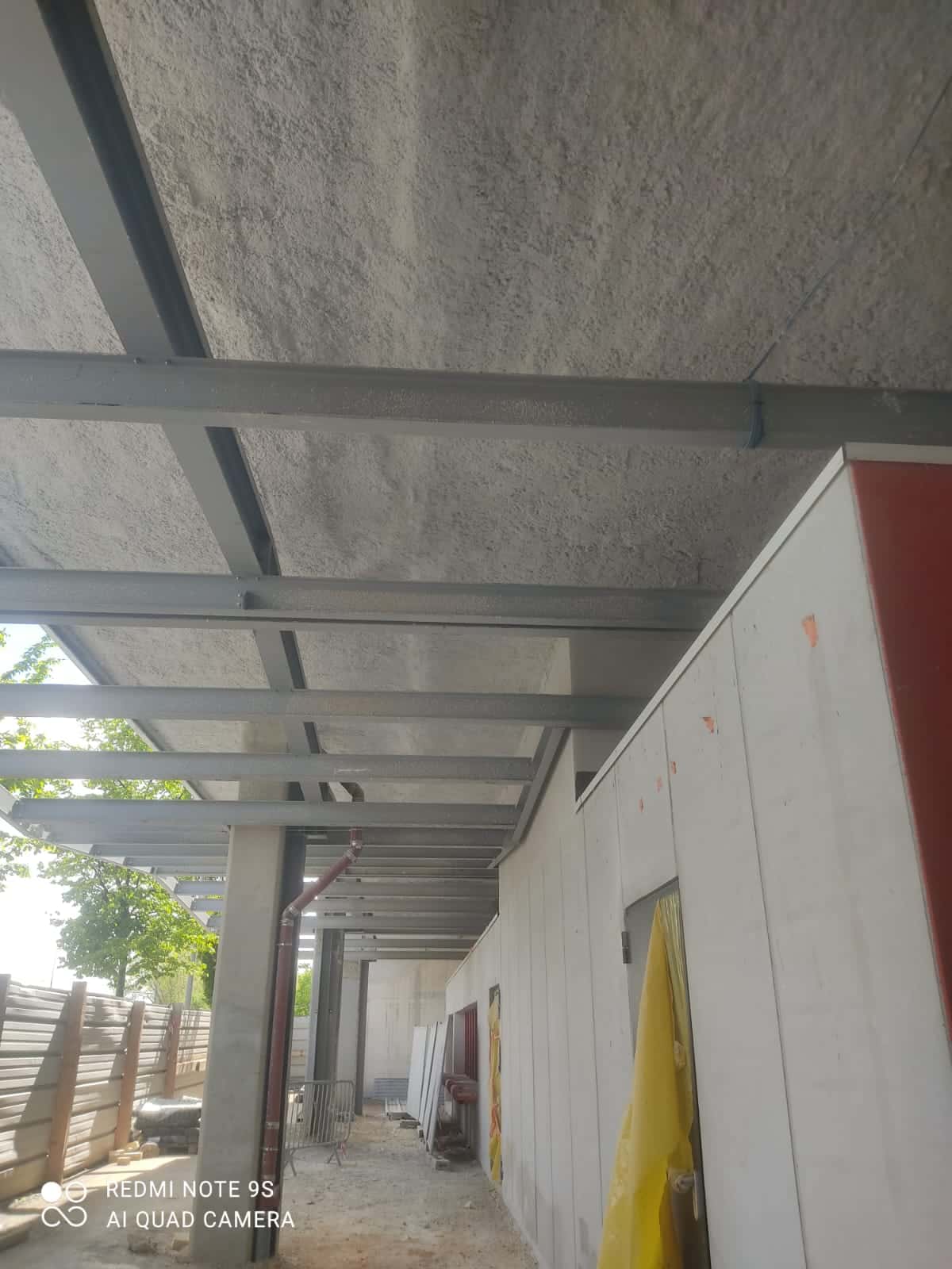 Structure métallique d'un porche de construction. Poutres grises sous un plafond texturé. Mur en panneaux isolants préfabriqués.