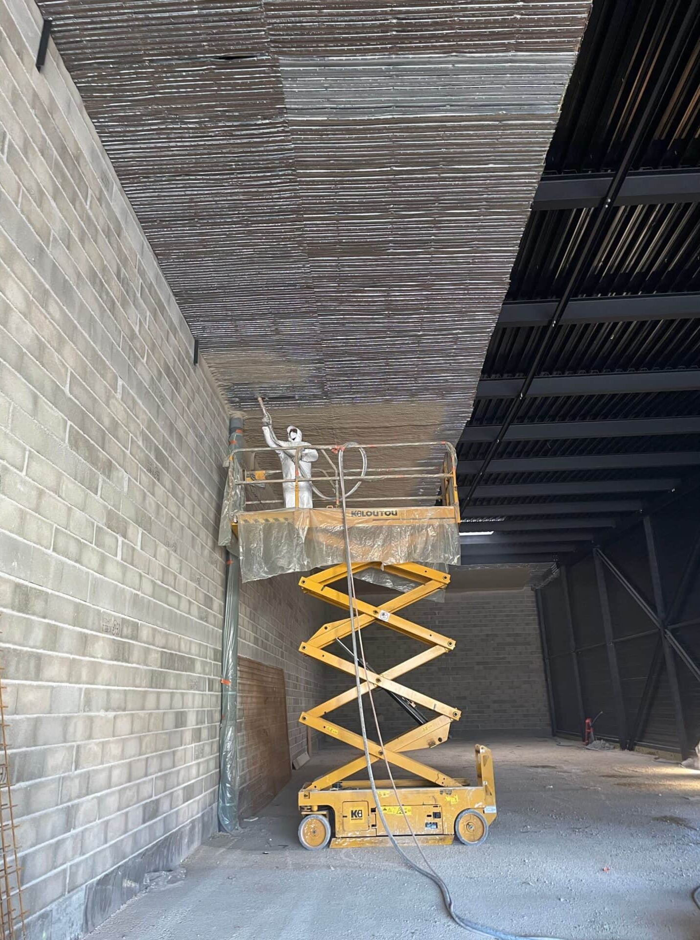Application Isolation Plafond Métallique Chantier Bâtiment Ouvrier en combinaison blanche appliquant un revêtement isolant sur un plafond métallique depuis une nacelle élévatrice jaune, près d'un mur en parpaings.