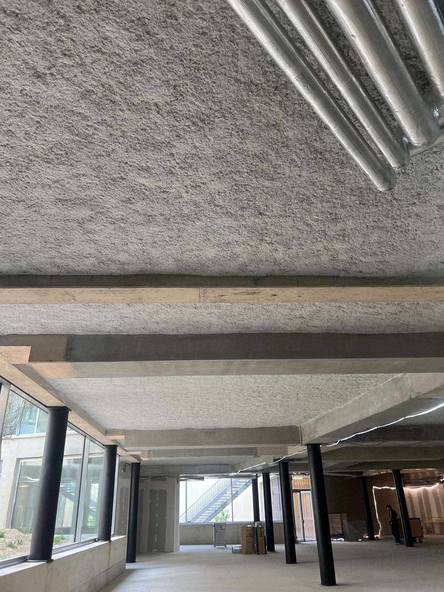 Flocage acoustique brut sur le plafond en béton d'un bâtiment moderne en construction, avec des poutres apparentes et des poteaux noirs cylindriques.