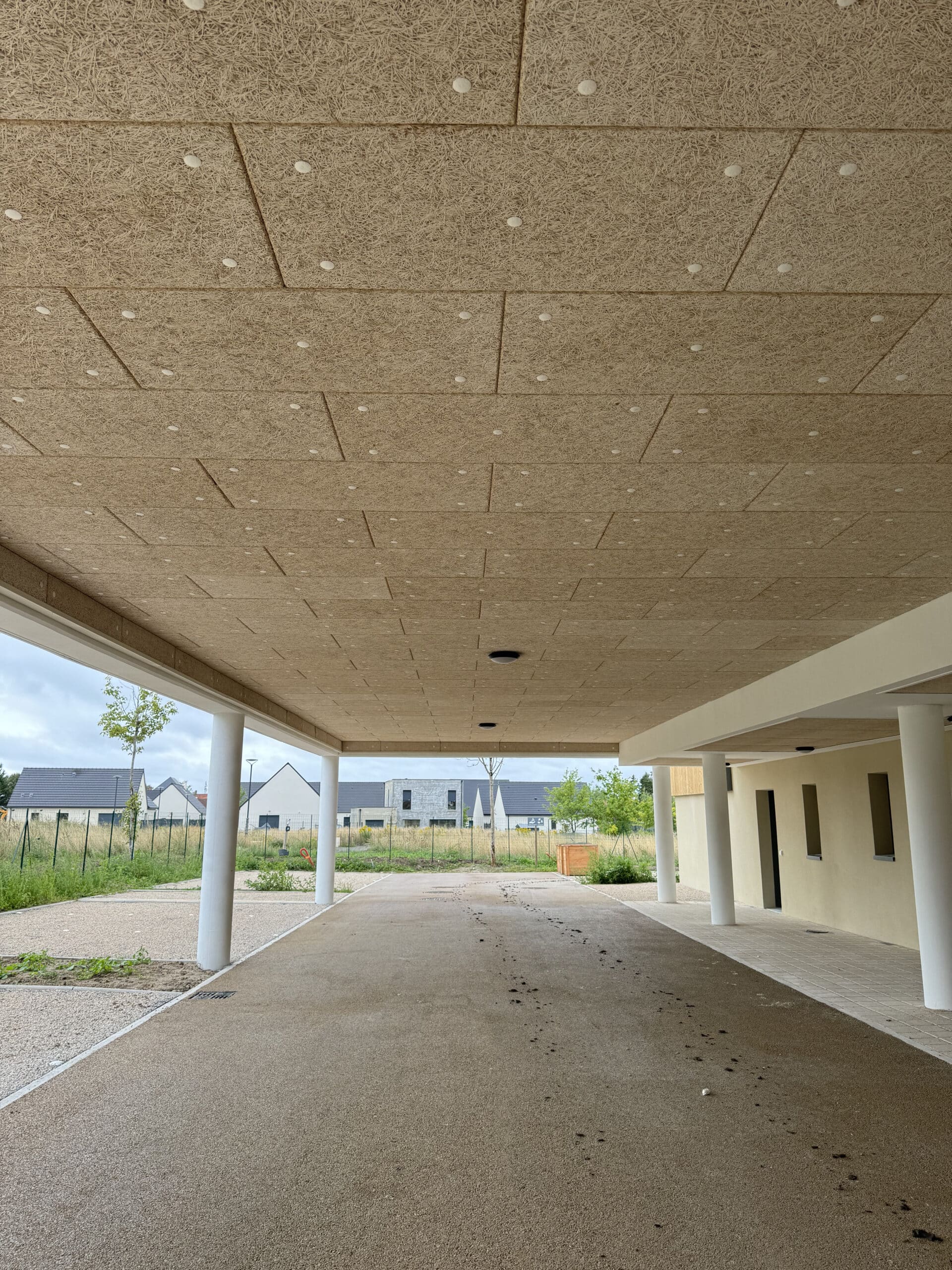 Entrée couverte moderne avec plafond acoustique en laine de bois. Allée en gravier résiné. Colonnes et façade jaune pâle. Lotissement neuf.