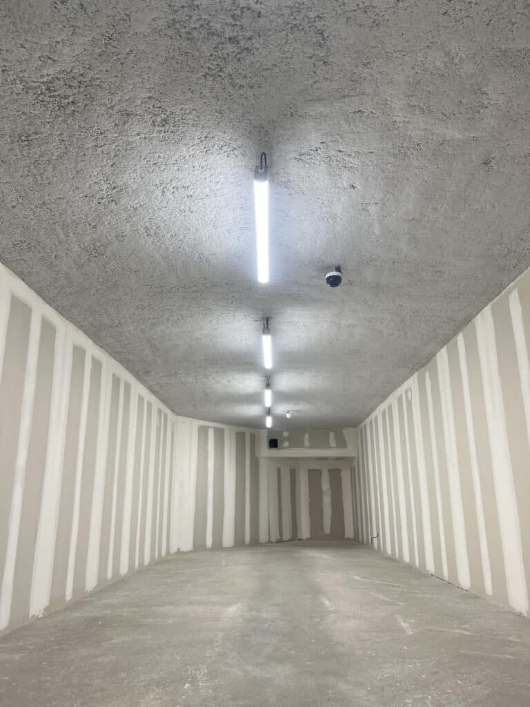 Couloir de chantier en perspective. Plafond béton brut, murs placo avec joints enduits. Lumières LED et caméra de surveillance.