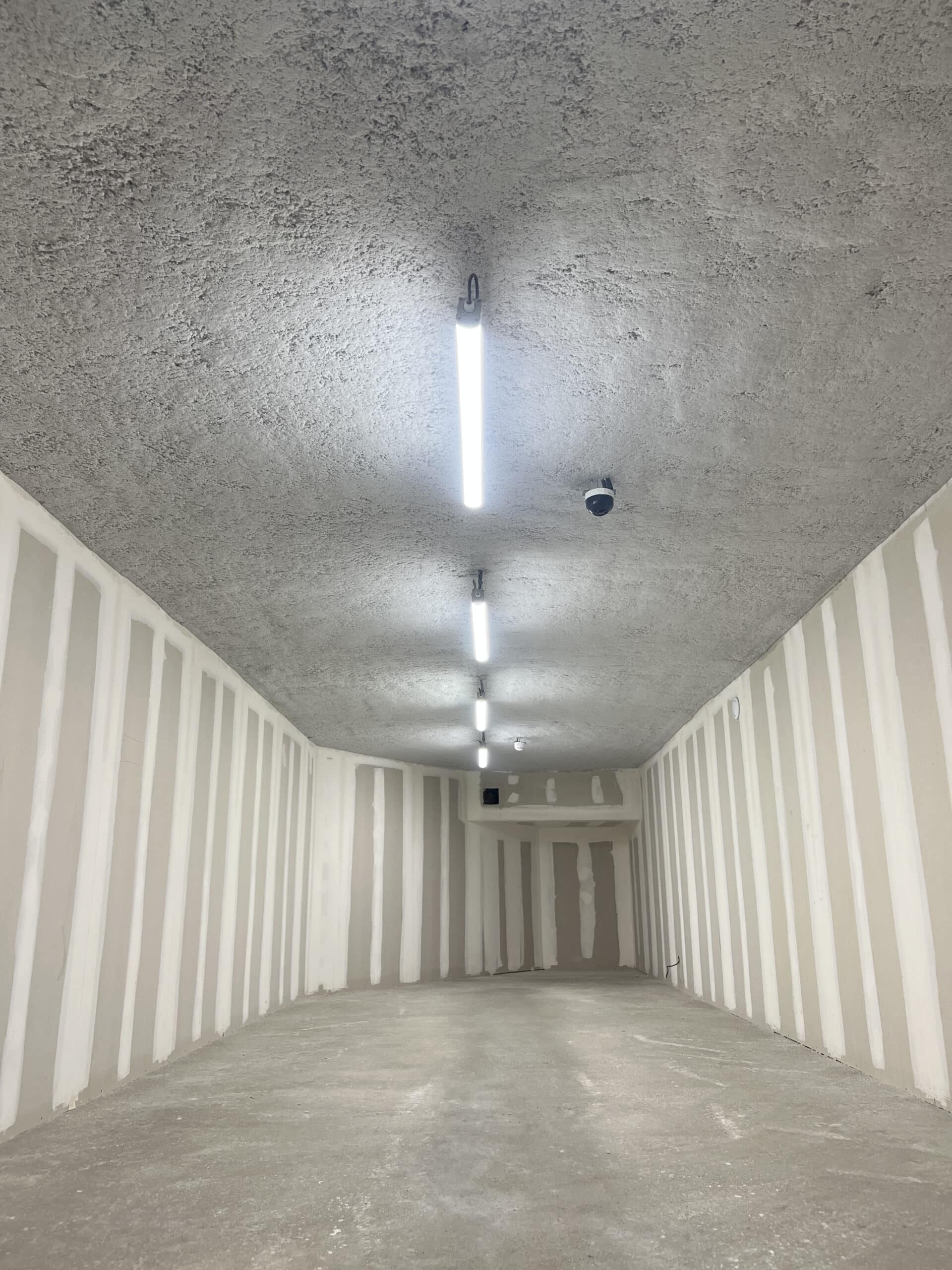Couloir de chantier en perspective. Plafond béton brut, murs placo avec joints enduits. Lumières LED et caméra de surveillance.