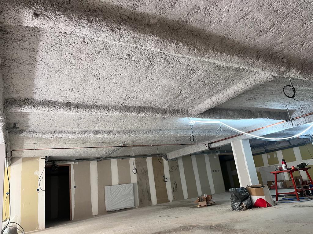 Plafond en béton recouvert d'isolation projetée (flocage) sur un chantier commercial, avec murs en placo et câbles exposés.