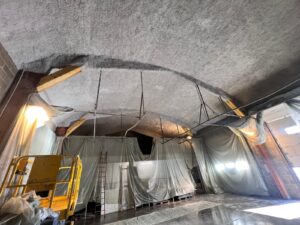 Isolation acoustique projetée sur un plafond voûté. Le chantier est protégé par des bâches de confinement et une nacelle est visible.